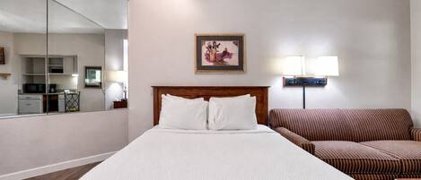 A hotel room with a large bed, a wooden headboard, a bedside table, a lamp, a couch, and a kitchen area visible through a glass partition.