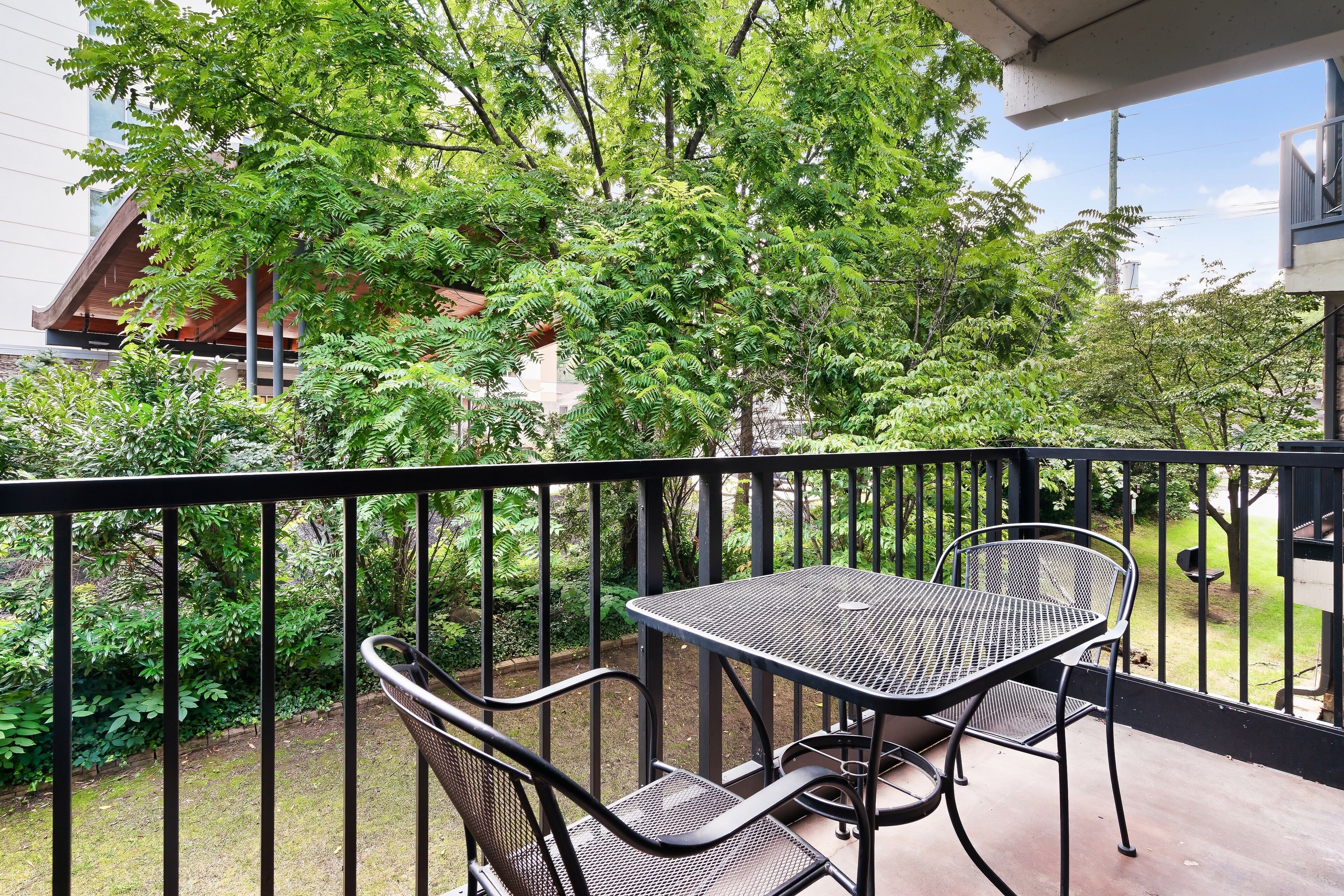 Ein Balkon mit Metalltisch und -stühlen, umgeben von Grünpflanzen und einem Gebäude im Hintergrund.