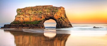 Formación natural de arco de roca con vista despejada al océano y un atardecer de fondo.