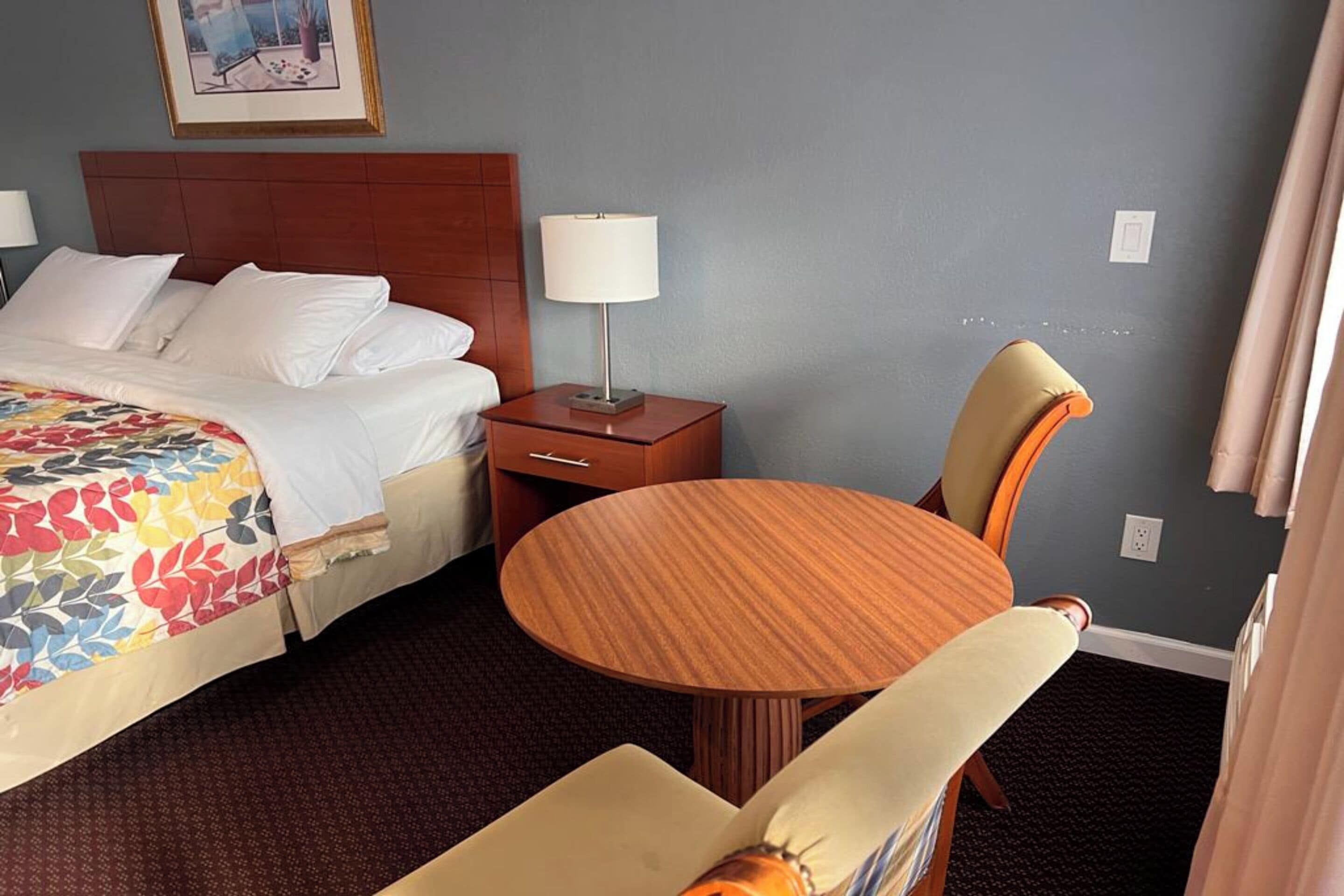 Quarto de hotel com cama, mesa de cabeceira com abajur, mesa redonda com cadeira e um quadro na parede.