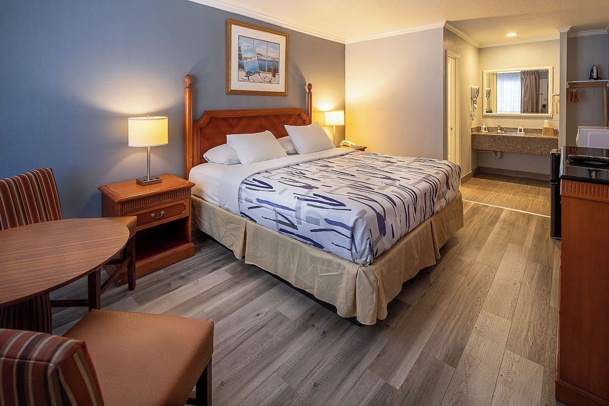 Quarto de hotel com cama, mesas de cabeceira, uma cadeira, uma mesinha, banheiro com pia e espelho, e um quadro na parede.