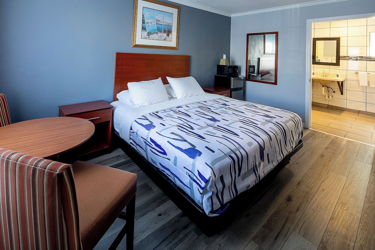 Quarto de hotel com uma cama, mesa de cabeceira de madeira, uma cadeira, uma mesa, um quadro na parede e um banheiro visível através de uma porta aberta.