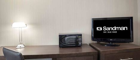 A hotel room with a desk, a microwave, a television, and a lamp.