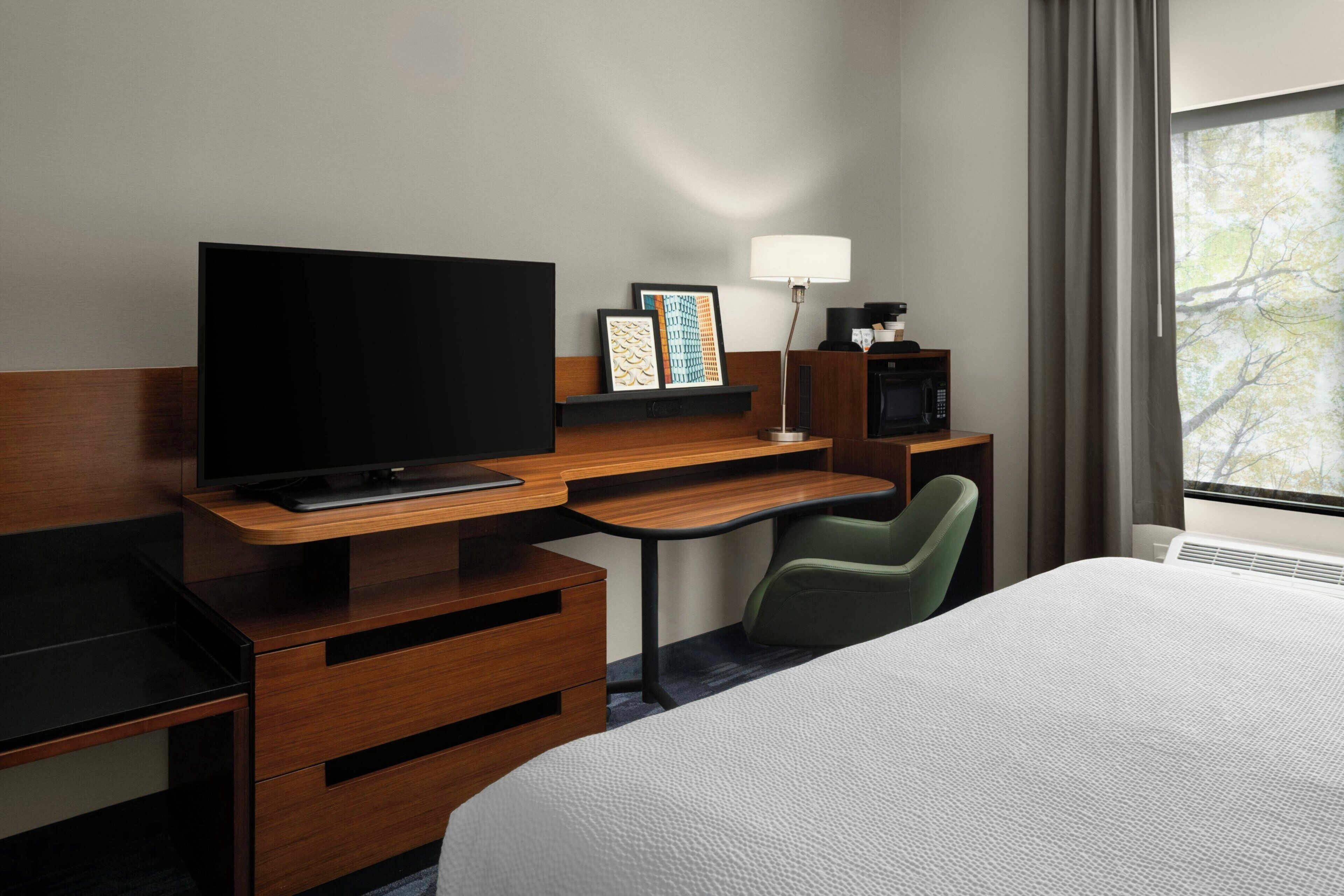 A hotel room with a bed, a desk with a TV, a chair, a lamp, and a microwave.