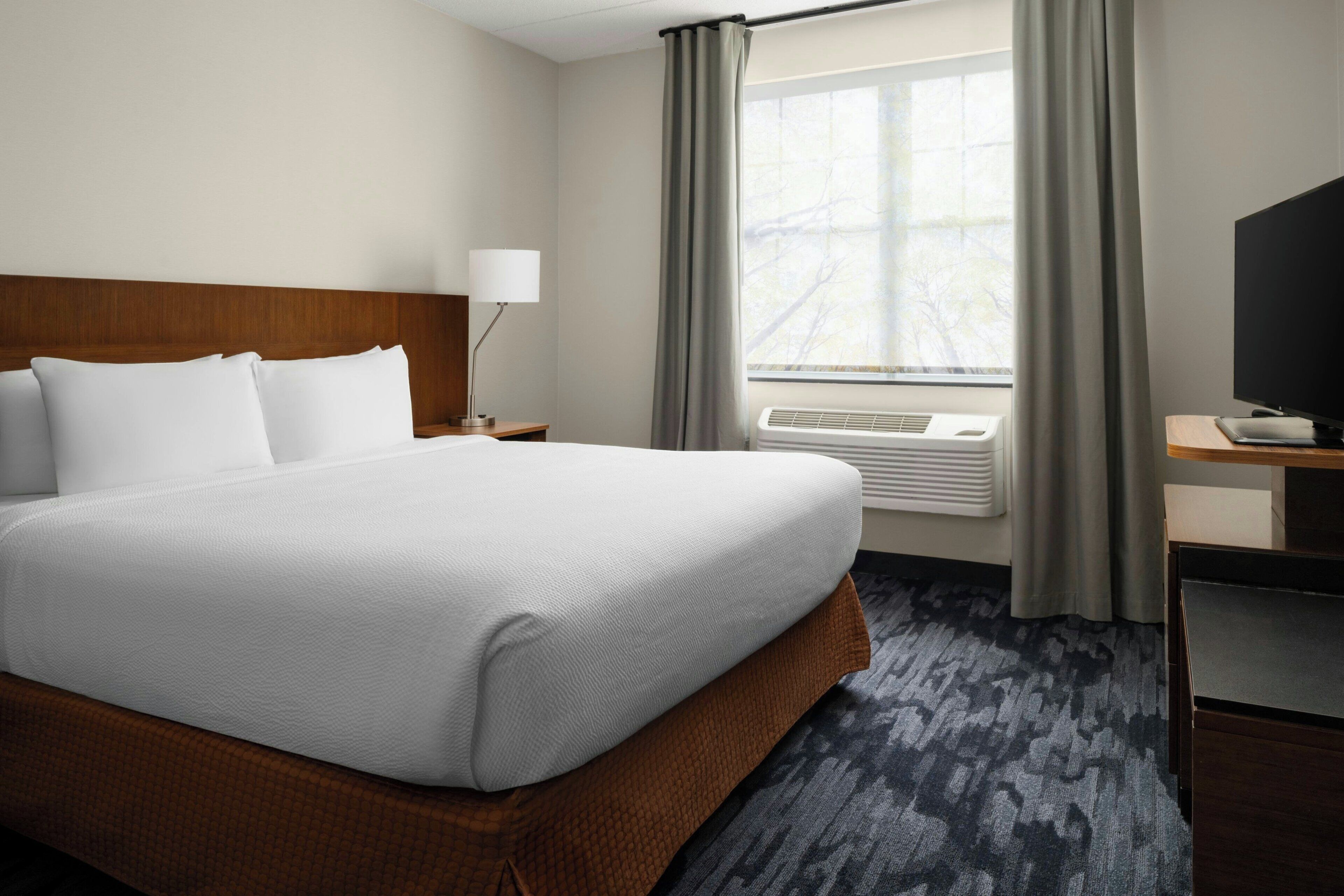 A hotel room with a large bed, a desk with a flat-screen TV, a window with curtains, and a wall-mounted air conditioner.