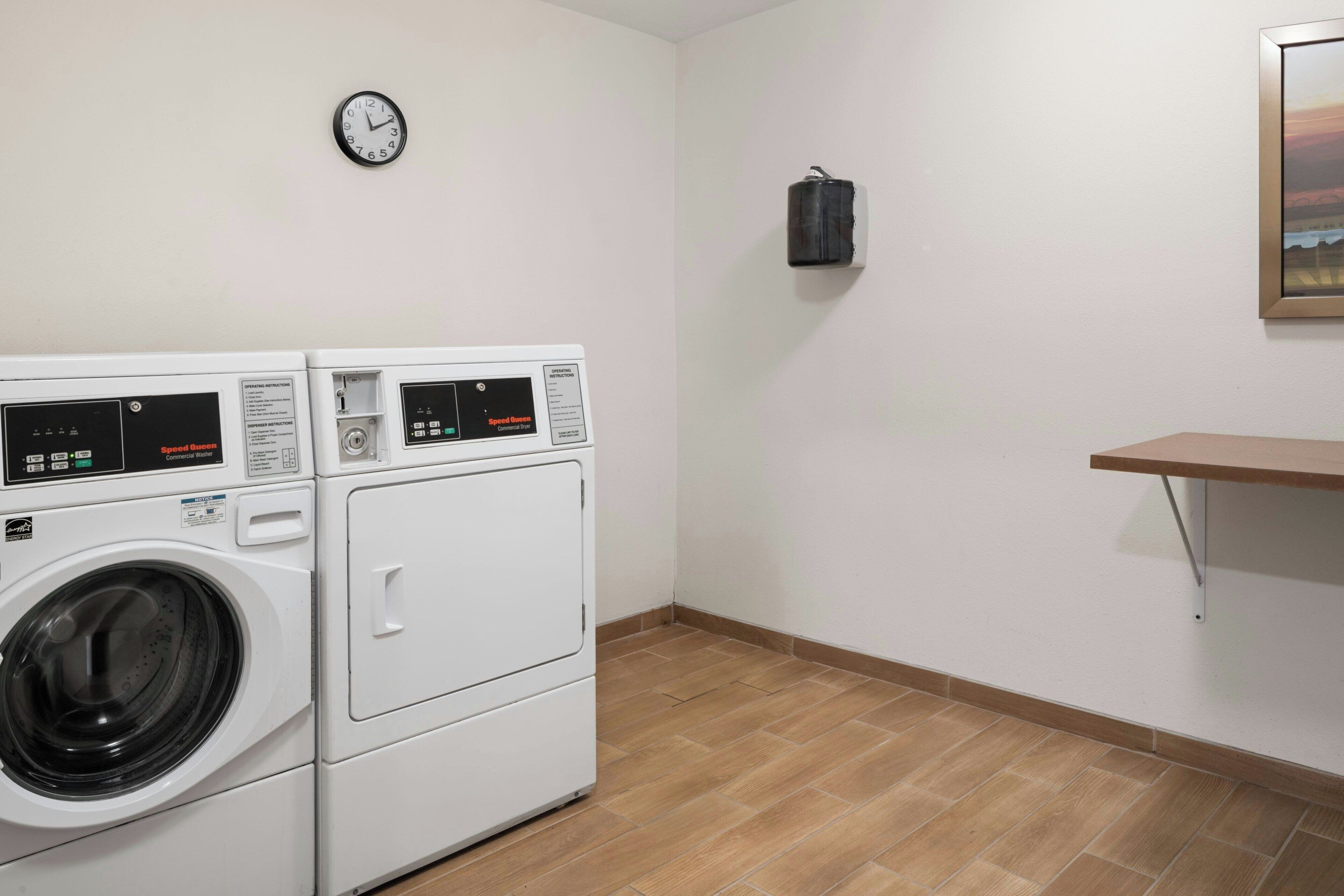 Un cuarto de lavandería con dos lavadoras apiladas, un reloj, un bote de basura negro y un estante de madera.