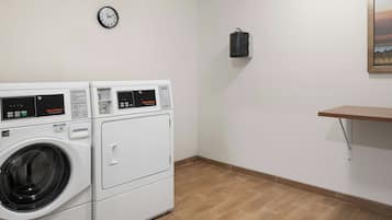 Un cuarto de lavandería con dos lavadoras apiladas, un reloj, un bote de basura negro y un estante de madera.