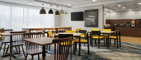 A hotel dining area with tables and chairs, a large window with curtains, a flat-screen TV, and a motivational poster on the wall.