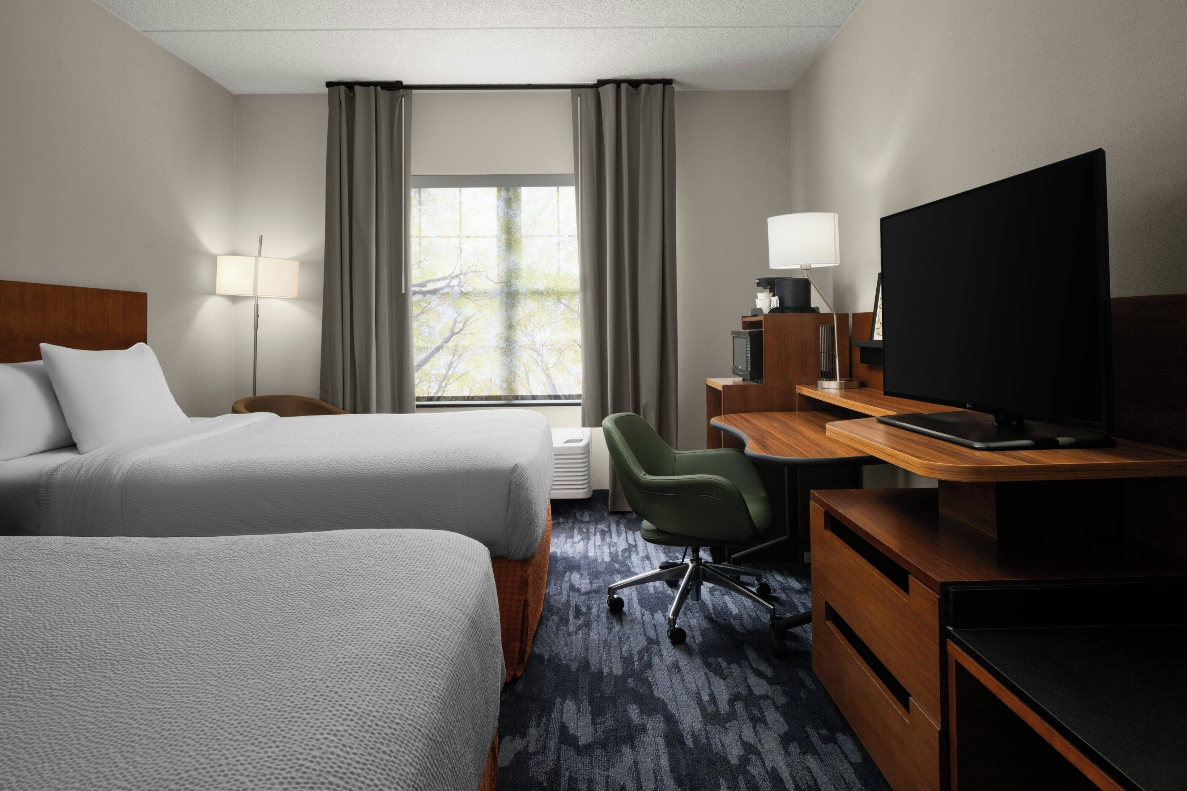 A hotel room with two beds, a desk with a computer, a television, and a window with curtains.