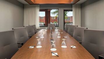 A conference room with a long table, grey chairs, and a view of a brick building through large windows.