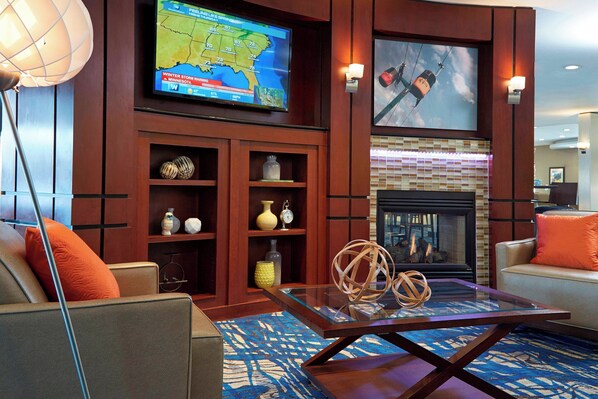 A modern living room with a fireplace, comfortable seating, a television displaying a weather map, and decorative items on shelves.