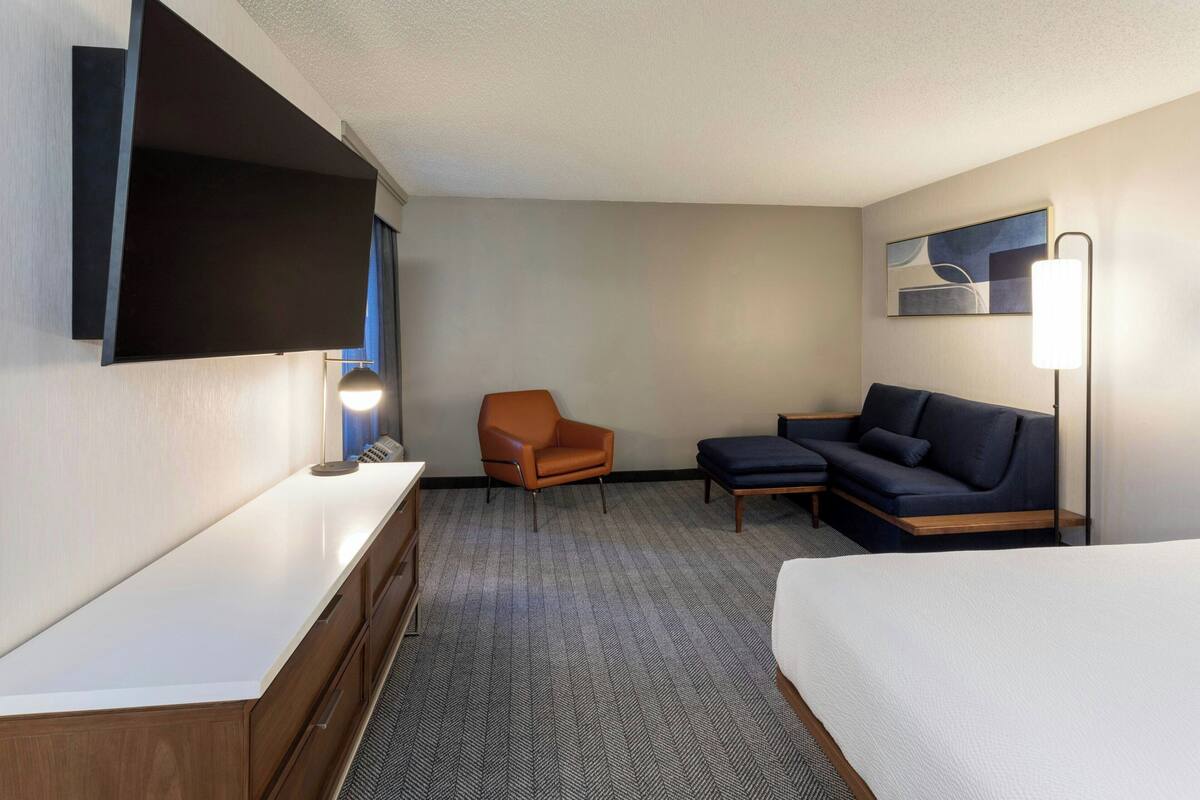 A hotel room with a flat-screen TV mounted on the wall, a wooden dresser, a brown armchair, a dark blue sofa, and a bedside table with a lamp.