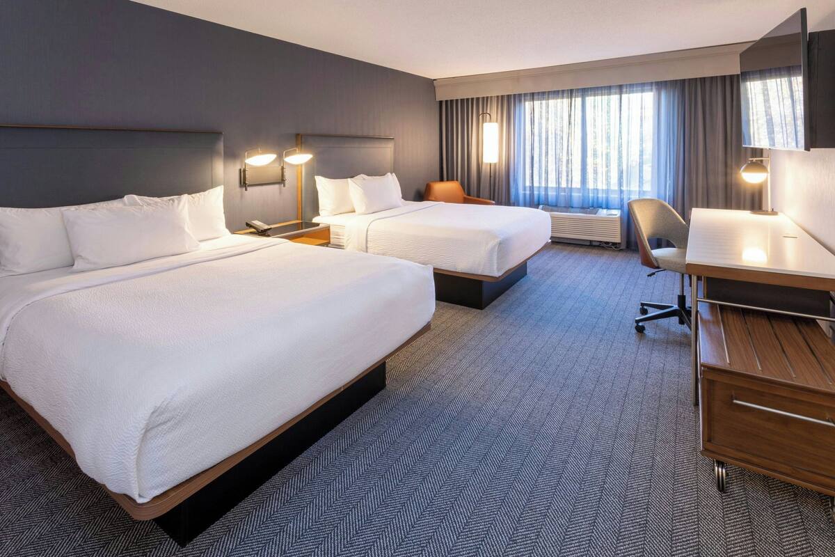 A hotel room with two beds, a desk, a chair, a desk lamp, and a large window with curtains.