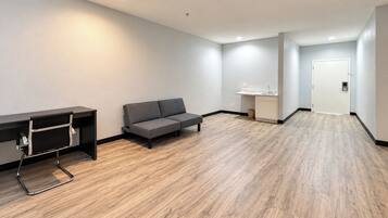 A modern, minimalistic room with a wooden floor, a grey sofa, a desk with a chair, and a small kitchenette area.