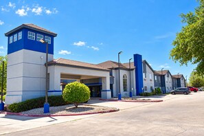 A hotel with a modern design, featuring a central entrance, a parking area, and a clear blue sky.