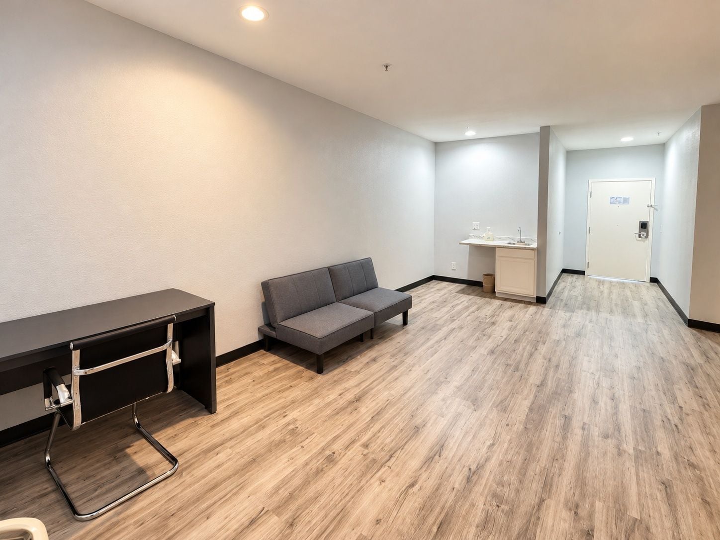 A hotel room with a desk, a sofa, a small kitchenette, and a door leading to another room.