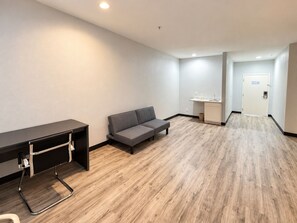 A hotel room with a desk, a sofa, a small kitchenette, and a door leading to another room.