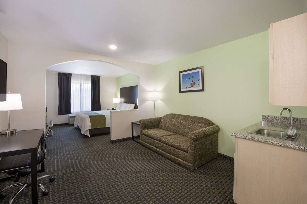 A hotel room with a bed, a sofa, a desk with a chair, a small kitchenette, and a bathroom.