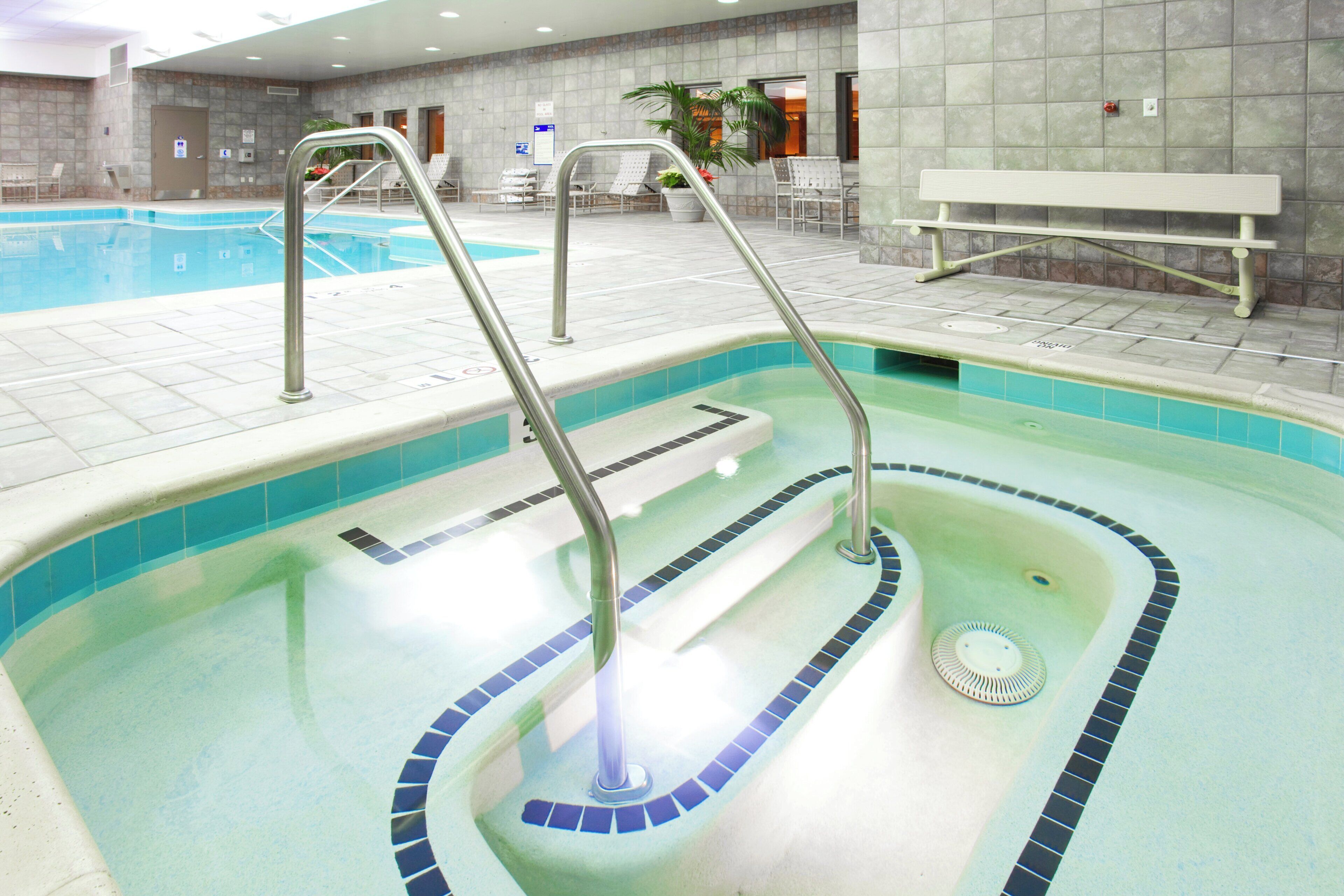 An indoor swimming pool with clear water, a diving board, and a lounge area with chairs and a bench.