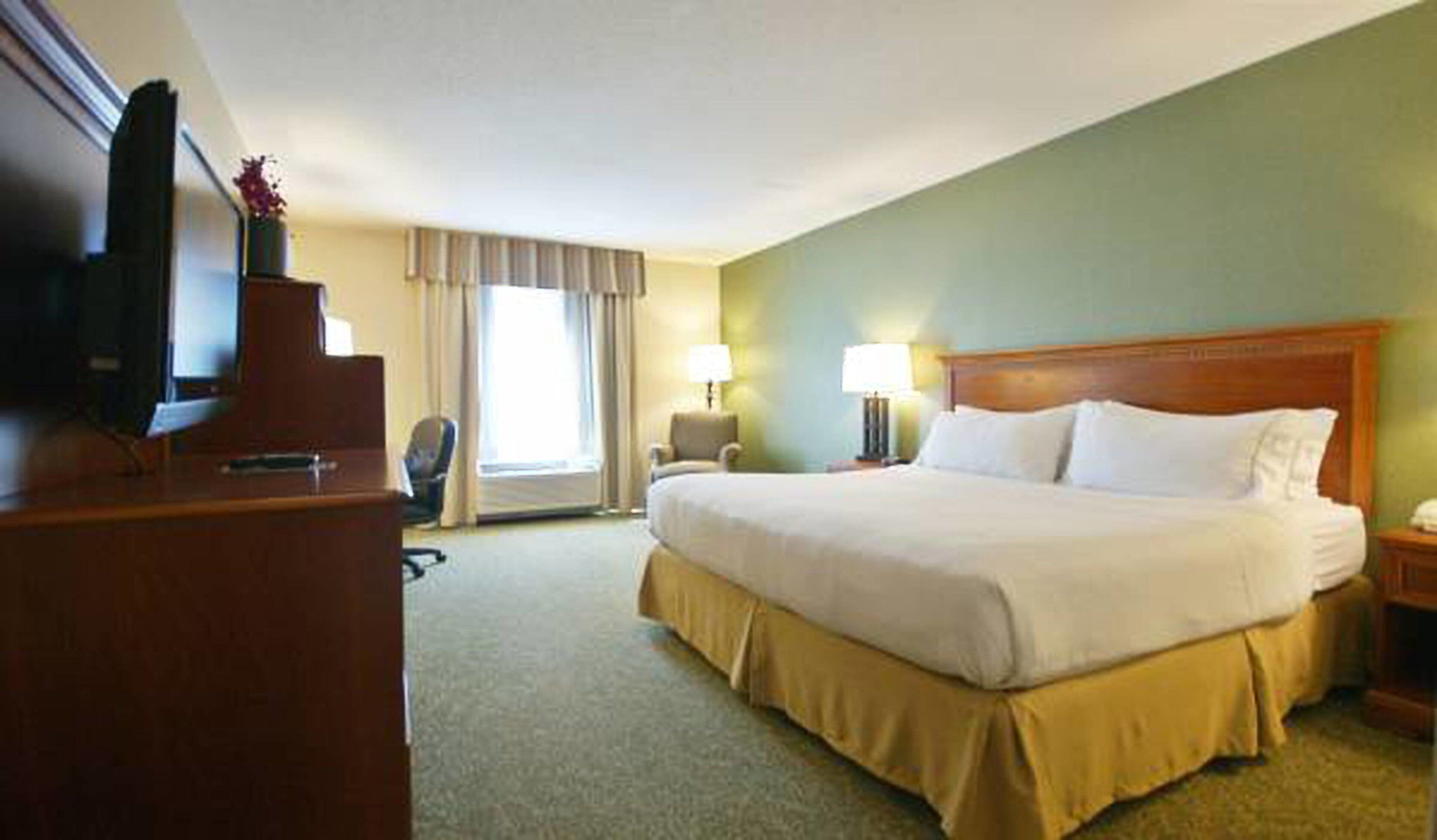 A hotel room with a large bed, a desk with a television, a chair, a lamp, and a window with curtains.