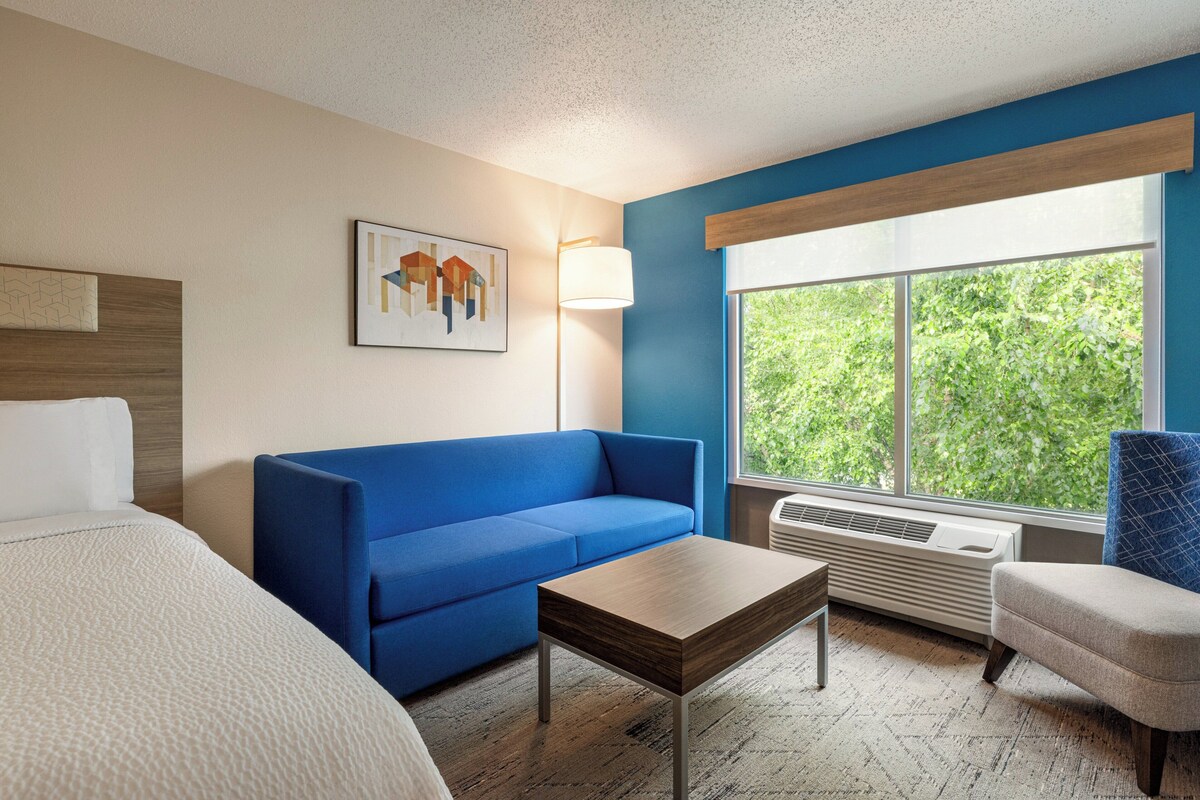 A hotel room with a blue sofa, a wooden coffee table, a bed with white bedding, a blue armchair, a floor lamp, and a large window with a view of greenery.