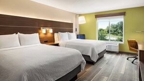 A hotel room with two beds, a desk, and a window with a view of greenery.