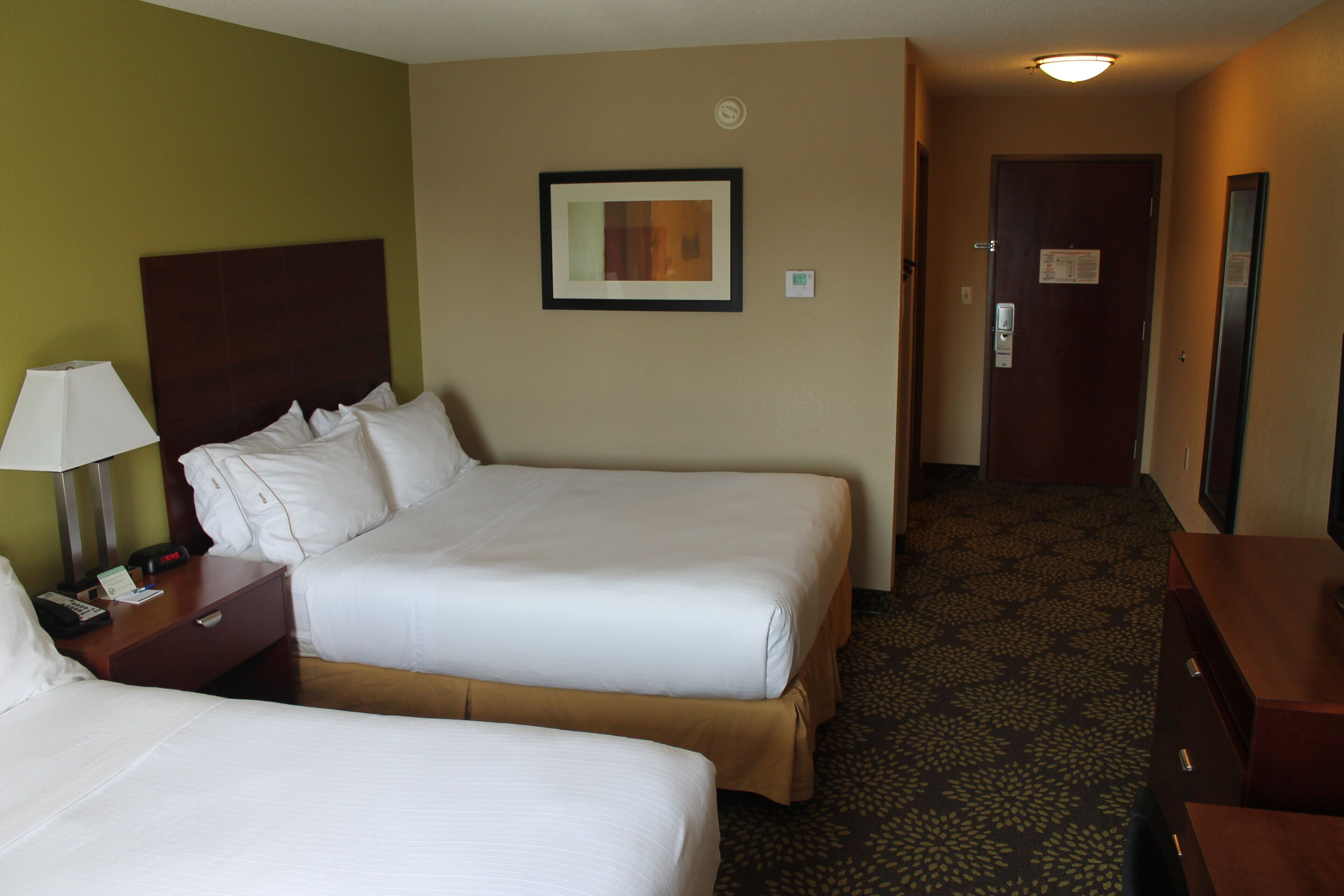 A hotel room with two beds, a nightstand with a lamp, a framed picture on the wall, and a carpeted floor.
