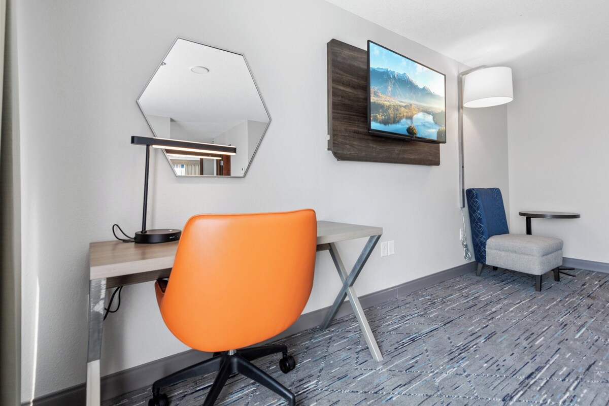 Camera d'albergo con scrivania, una sedia arancione, un tavolino, una lampada e TV a parete.