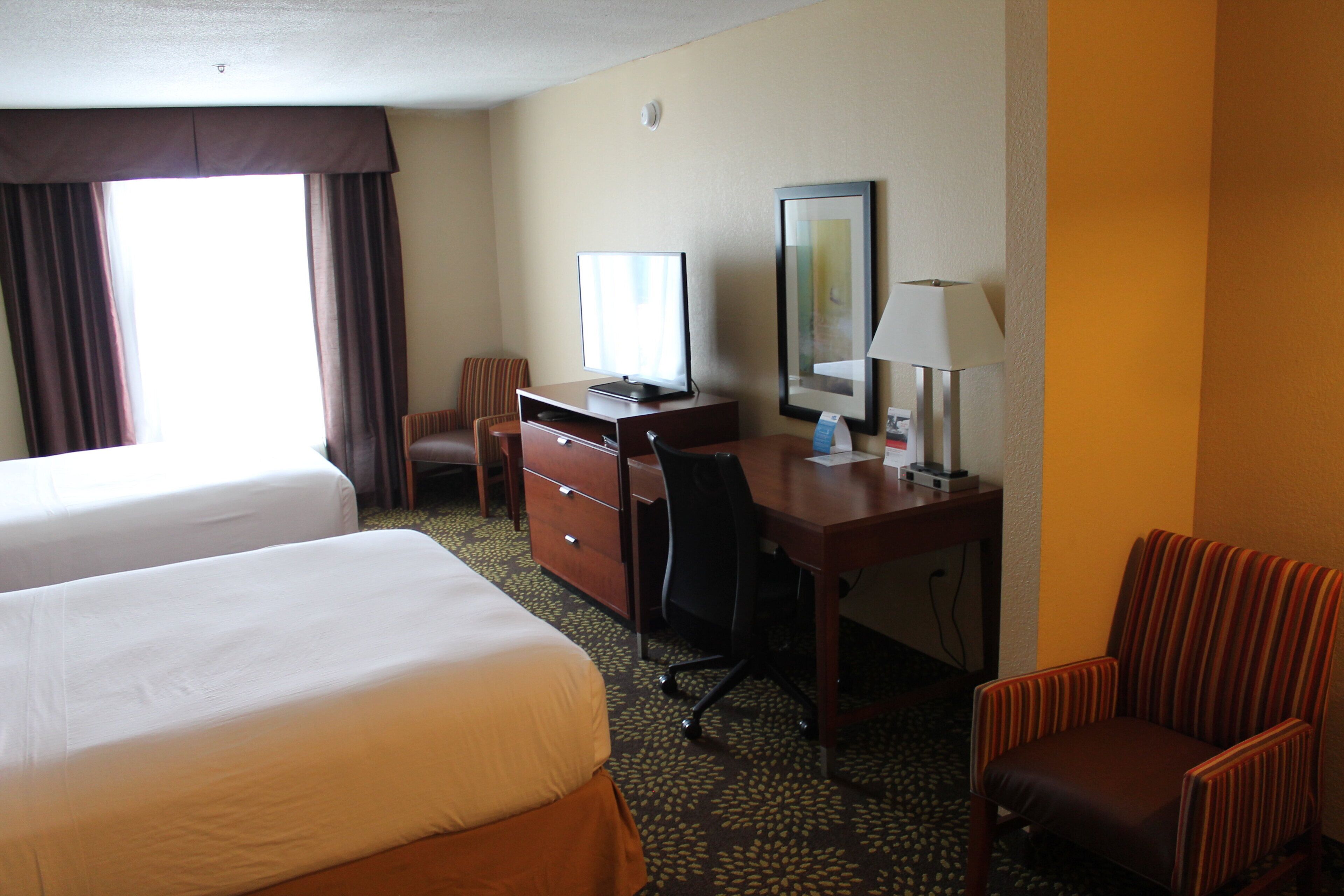 Een hotelkamer met twee bedden, een bureau met een computer, een stoel, een lamp en een raam met gordijnen.