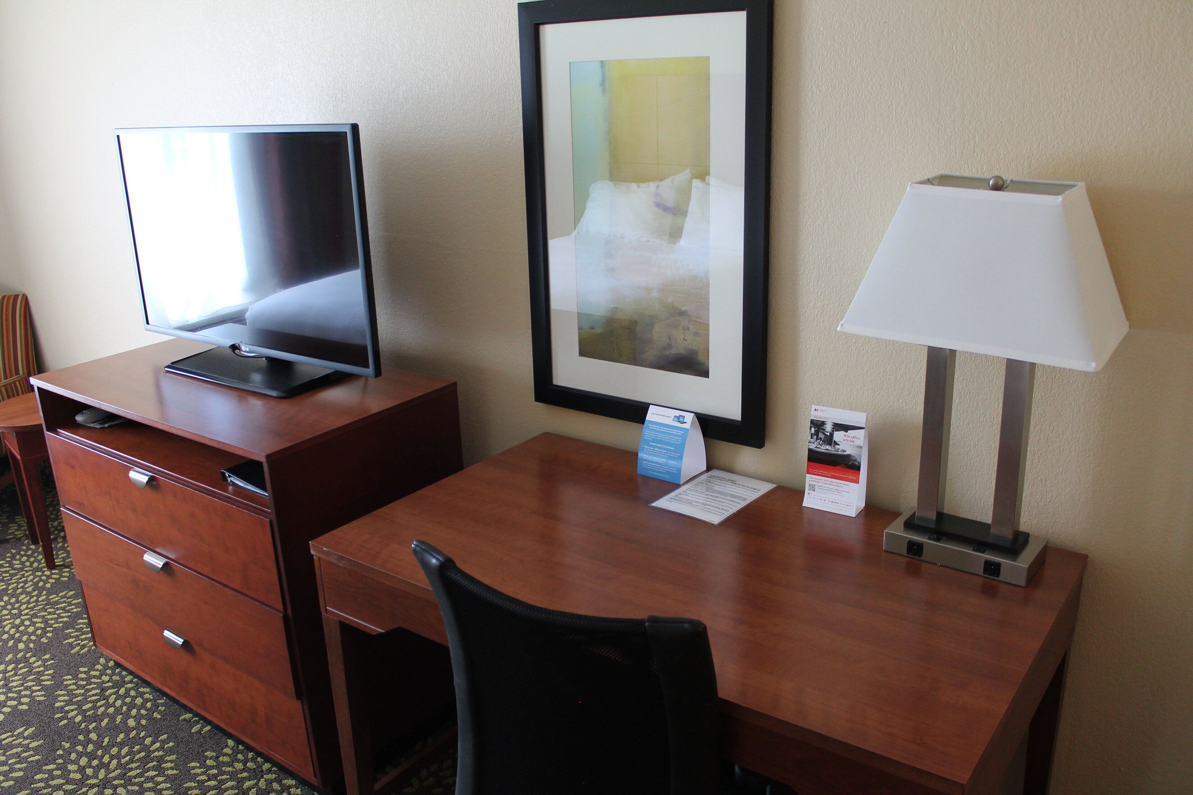 A hotel room with a desk, a chair, a TV, a framed picture on the wall, and a lamp.