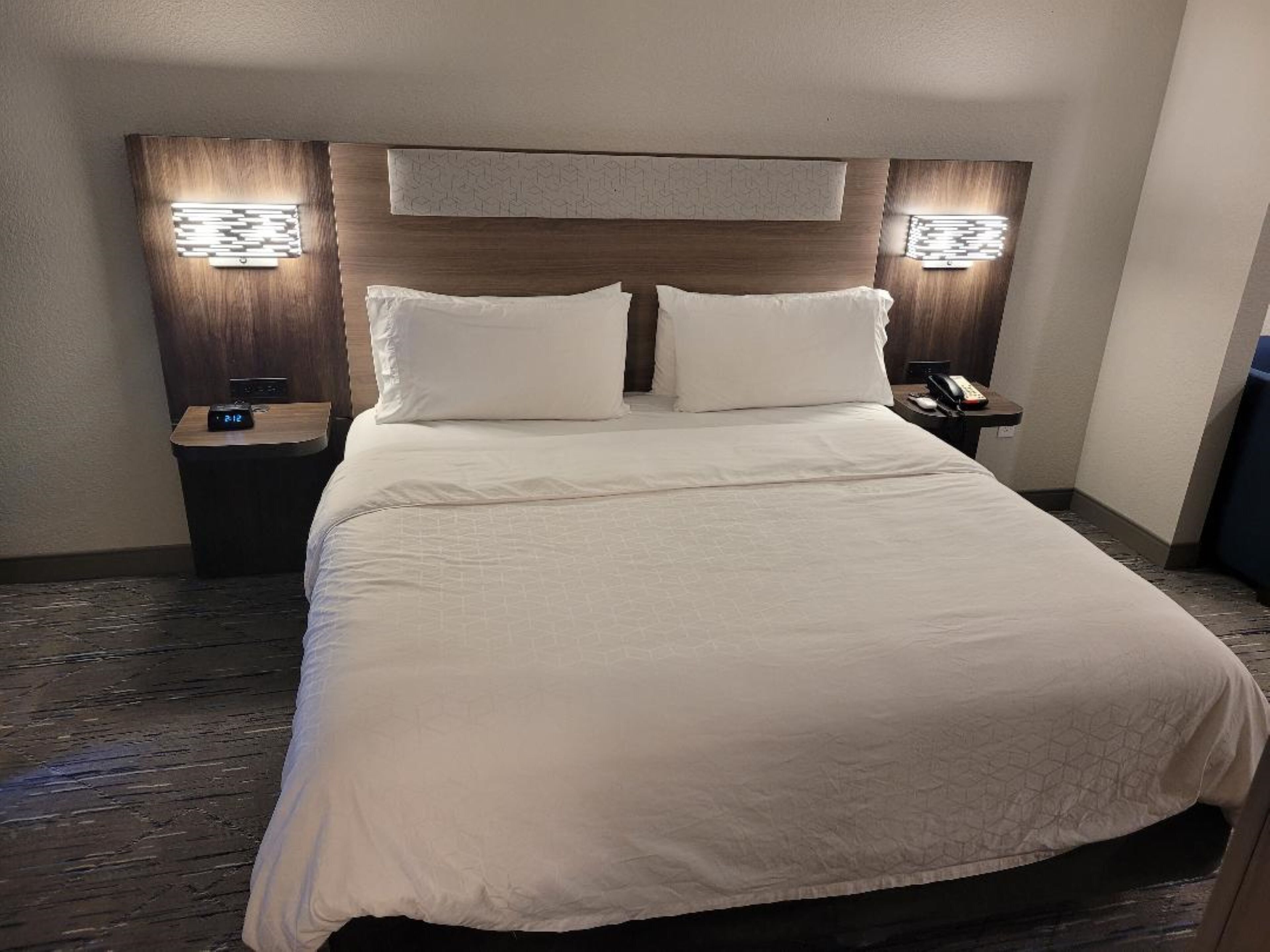 A hotel room with a large bed, two bedside tables, a nightstand, and wall-mounted lighting.