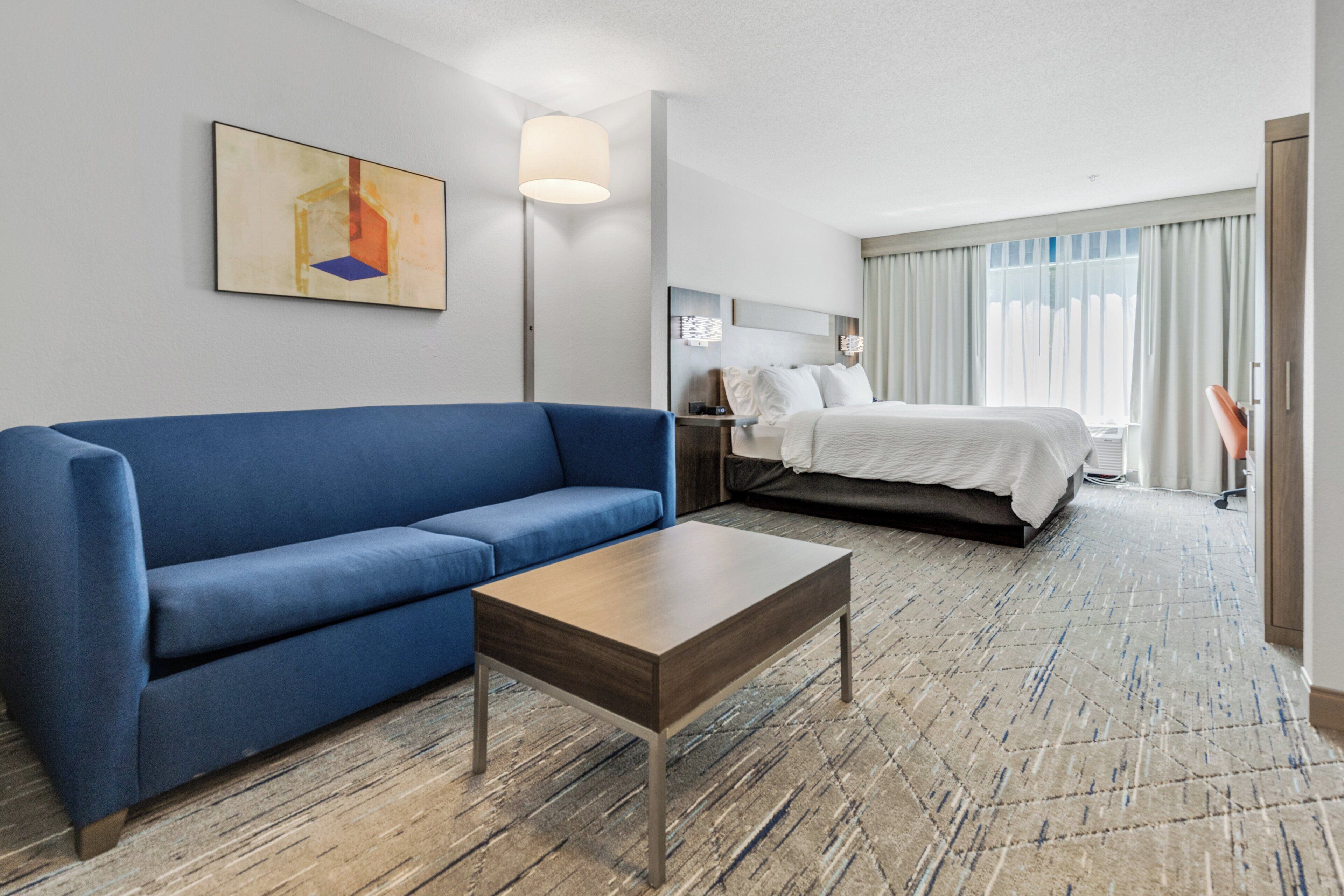 A hotel room with a blue sofa, a wooden coffee table, a bed with white bedding, a nightstand, and a painting on the wall.