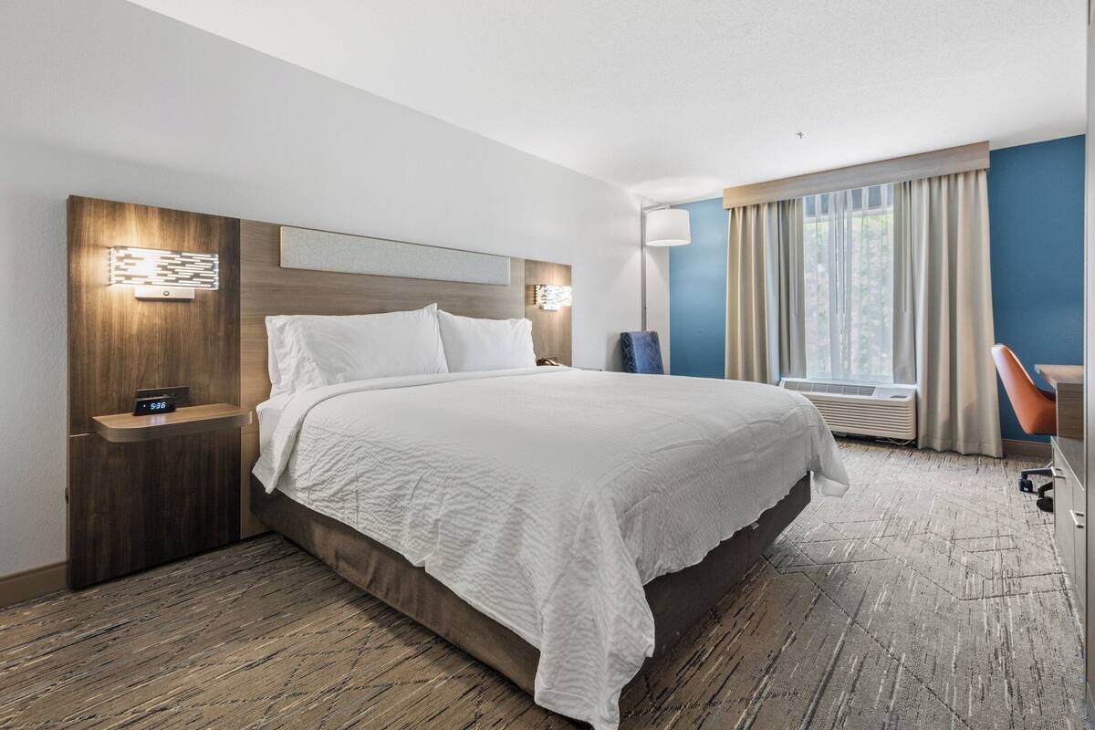 A hotel room with a large bed, a wooden headboard, a nightstand with a clock, a desk with a chair, and a window with curtains.