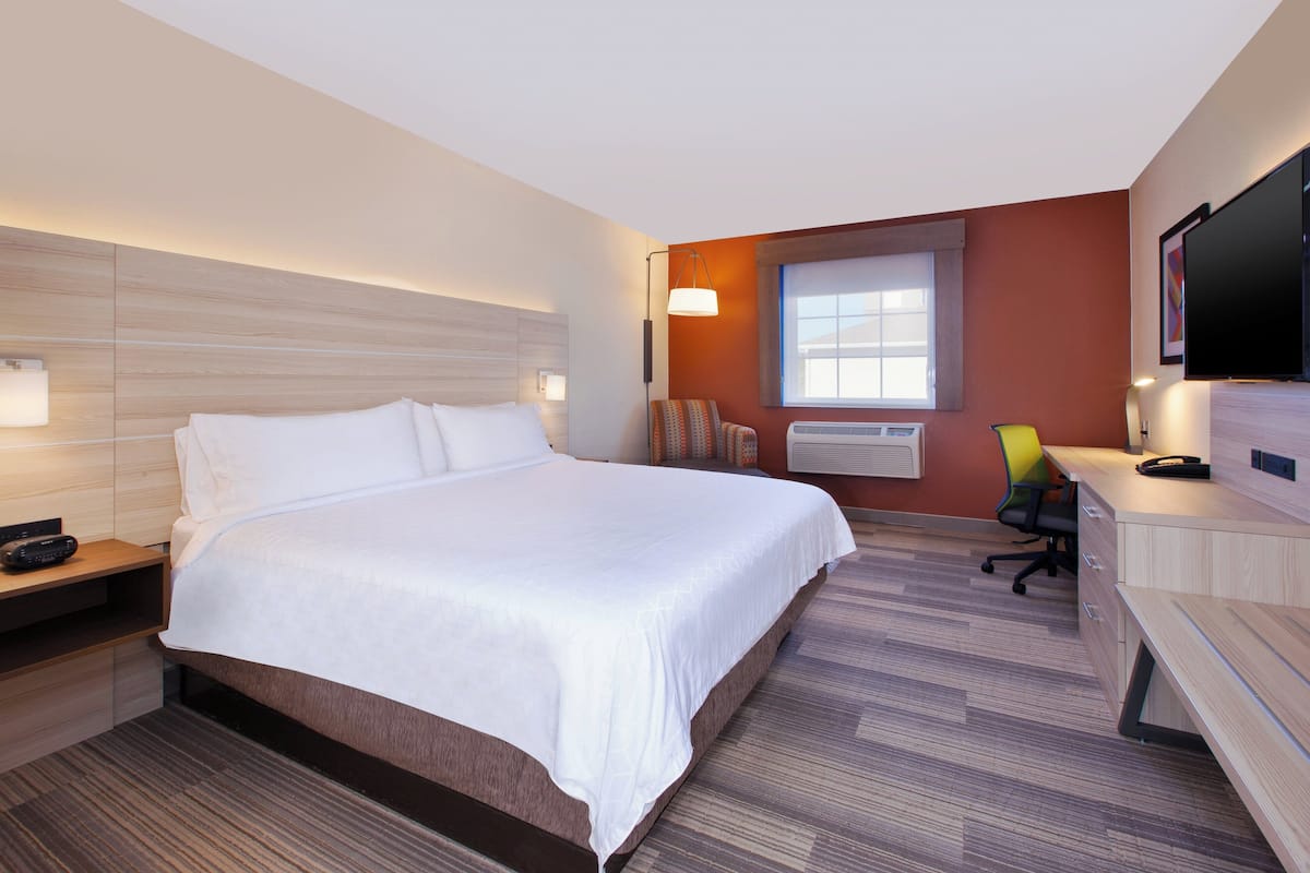 A hotel room with a large bed, a desk with a chair, a flat-screen TV, a window, and a wall-mounted air conditioning unit.