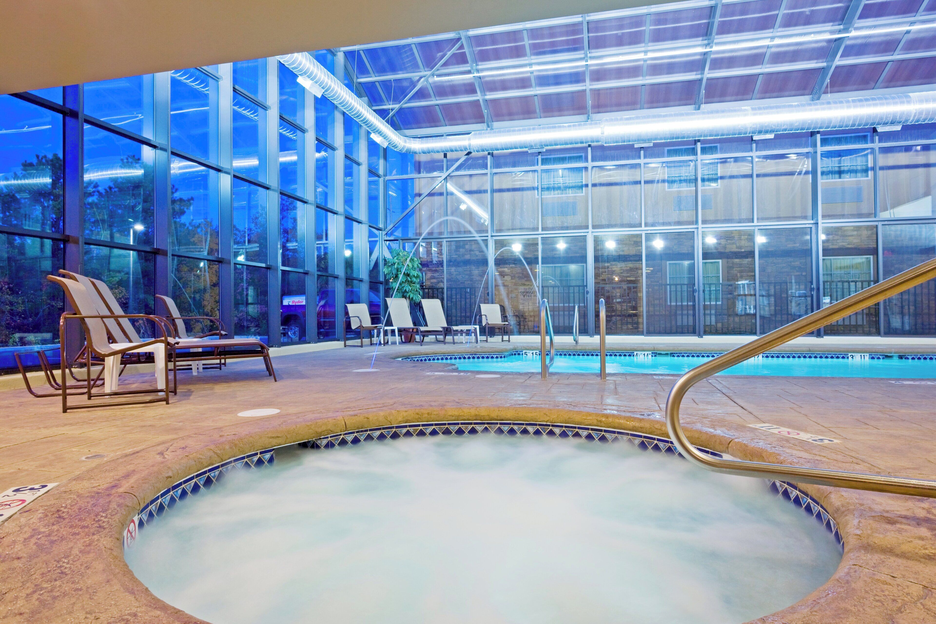 An indoor swimming pool with a glass enclosure, lounge chairs, and a hot tub.