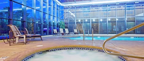 An indoor swimming pool with a glass enclosure, lounge chairs, and a hot tub.