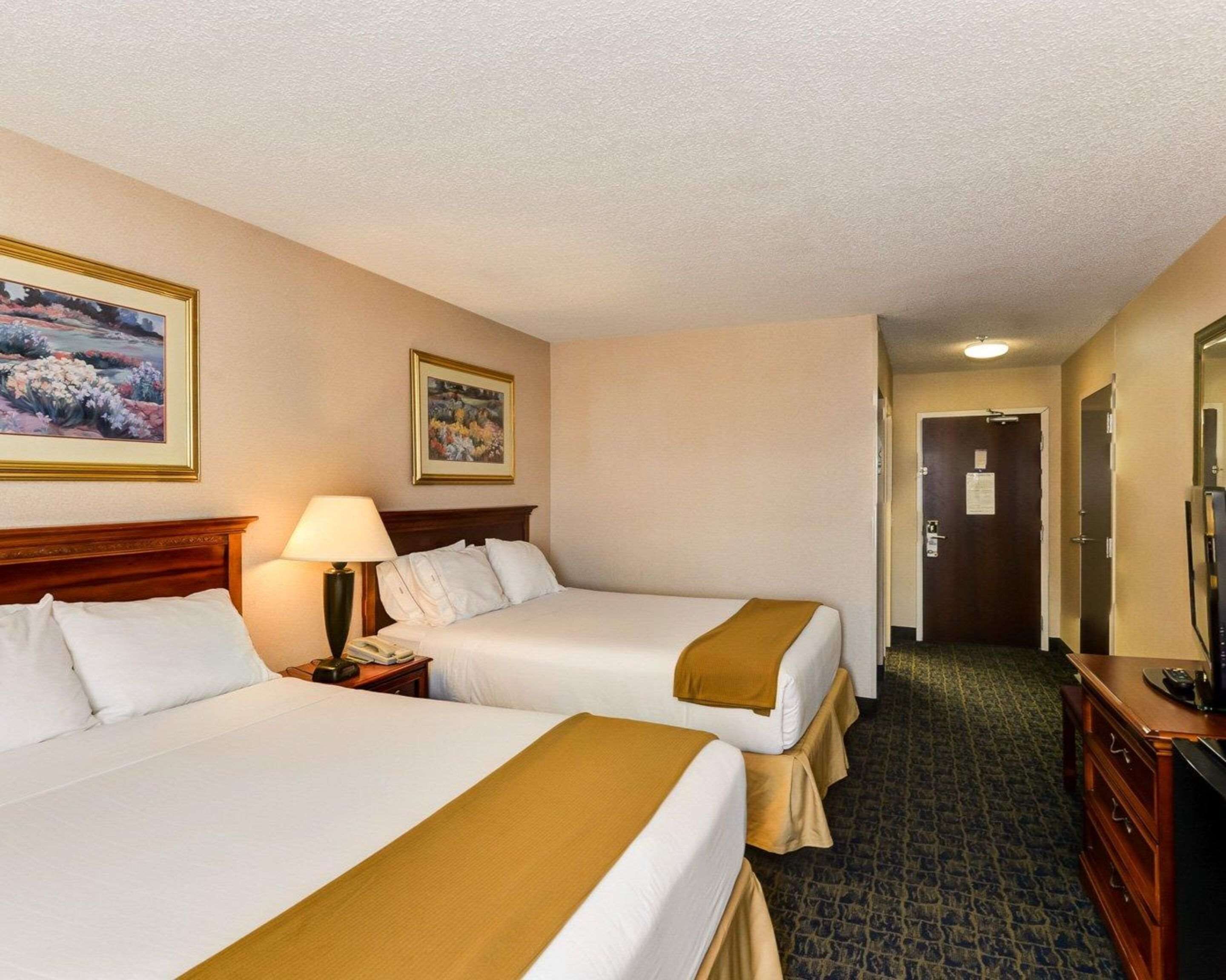 A hotel room with two beds, a desk, a lamp, a TV, and a door leading to a hallway.