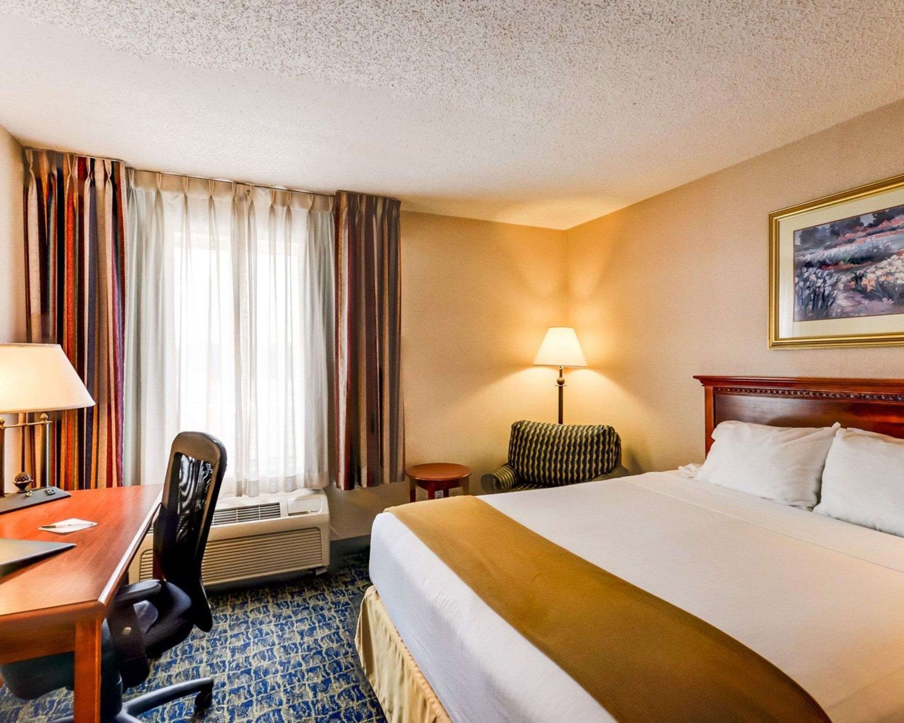 Quarto de hotel com cama, escrivaninha com cadeira, luminária e um quadro na parede.