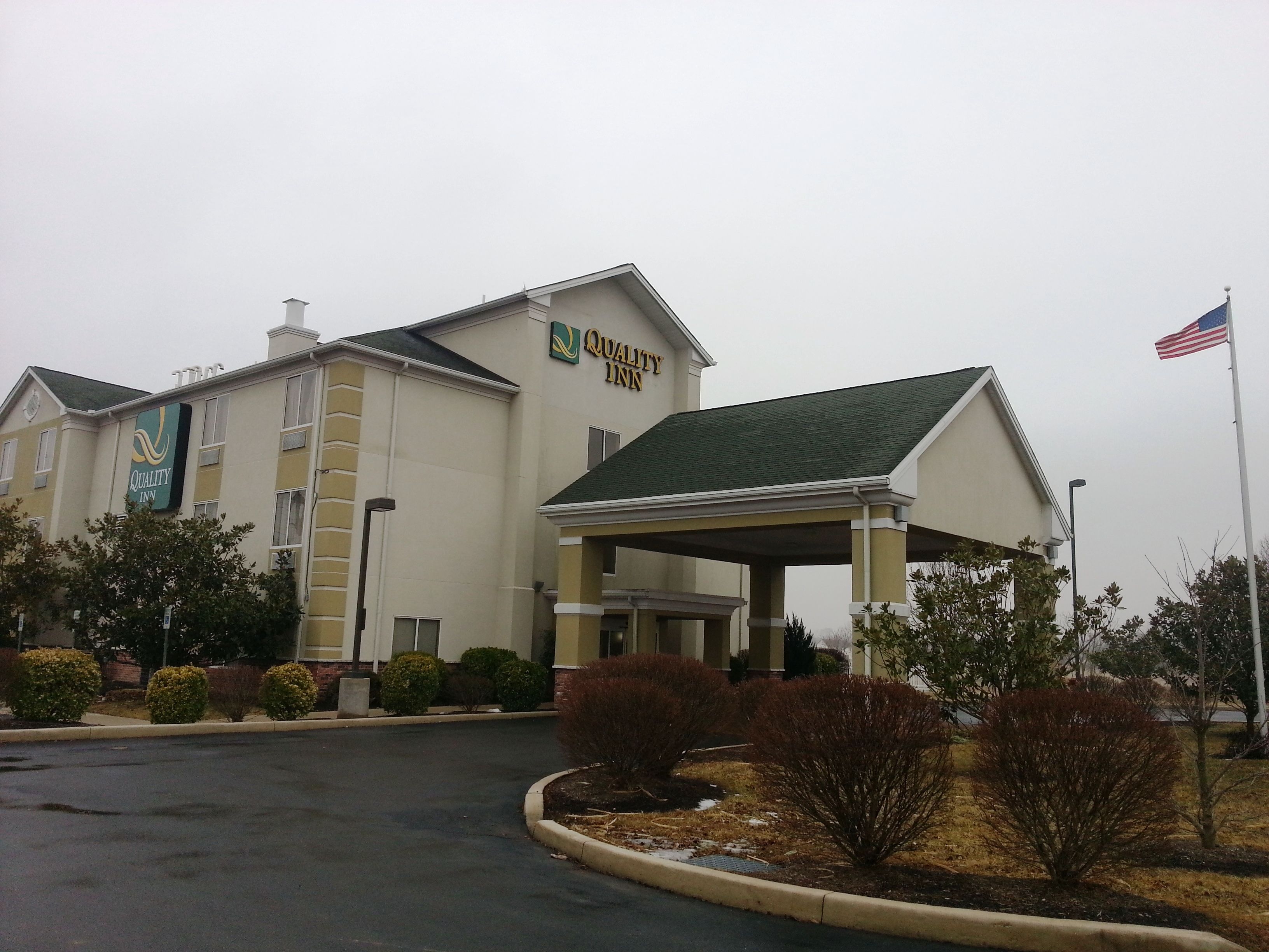 A Quality Inn hotel with a covered entrance, American flag, and landscaped area with shrubs.