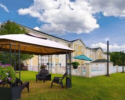 Un área de estar al aire libre techada con sillas y sombrillas, una piscina y un edificio con cerca blanca al fondo.