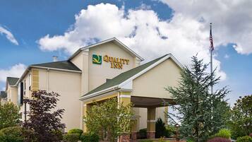 Un hotel Quality Inn con fachada color beige, logotipo verde y amarillo, y un césped delantero bien cuidado con diversos arbustos e izado.