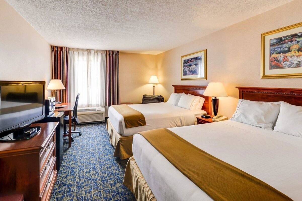 Ein Hotelzimmer mit zwei Betten, einem Schreibtisch mit Fernseher und einem großen Fenster mit Vorhängen.
