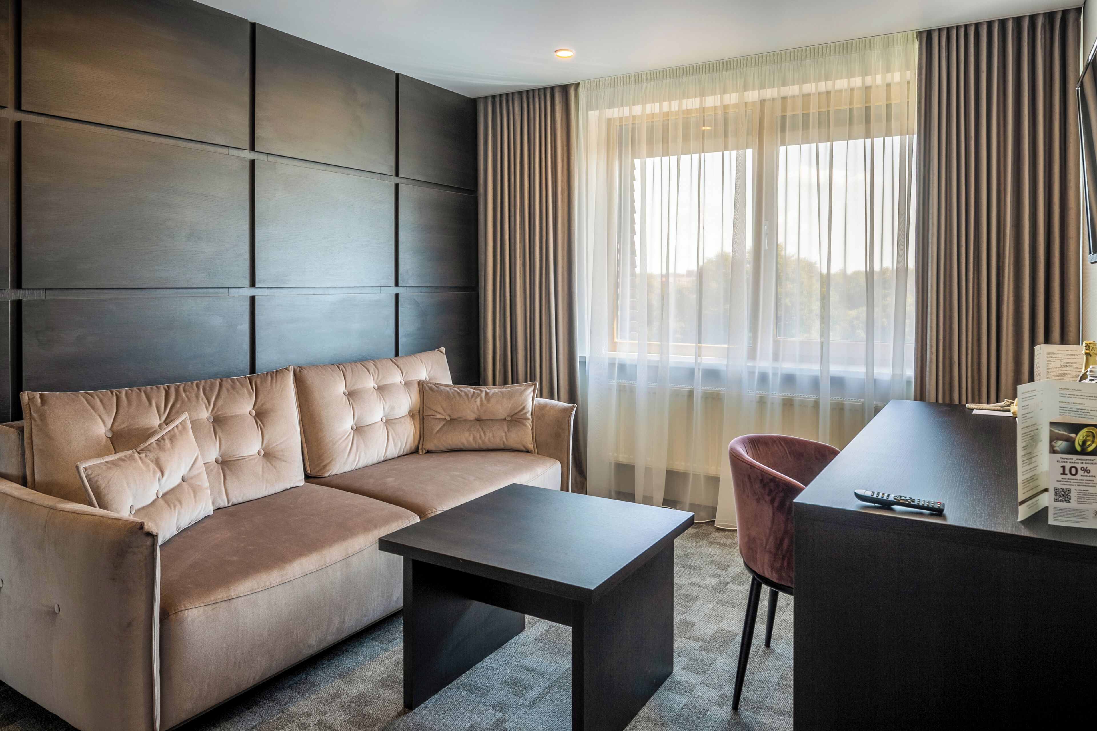 A modern hotel room with a sofa, a small table, a chair, and a desk with a remote control.