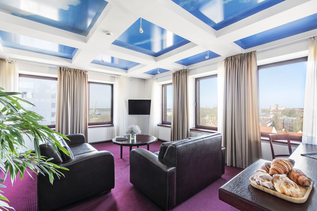 A modern hotel room with a large window, a flat-screen TV, a sofa, a dining table with pastries, and a potted plant.