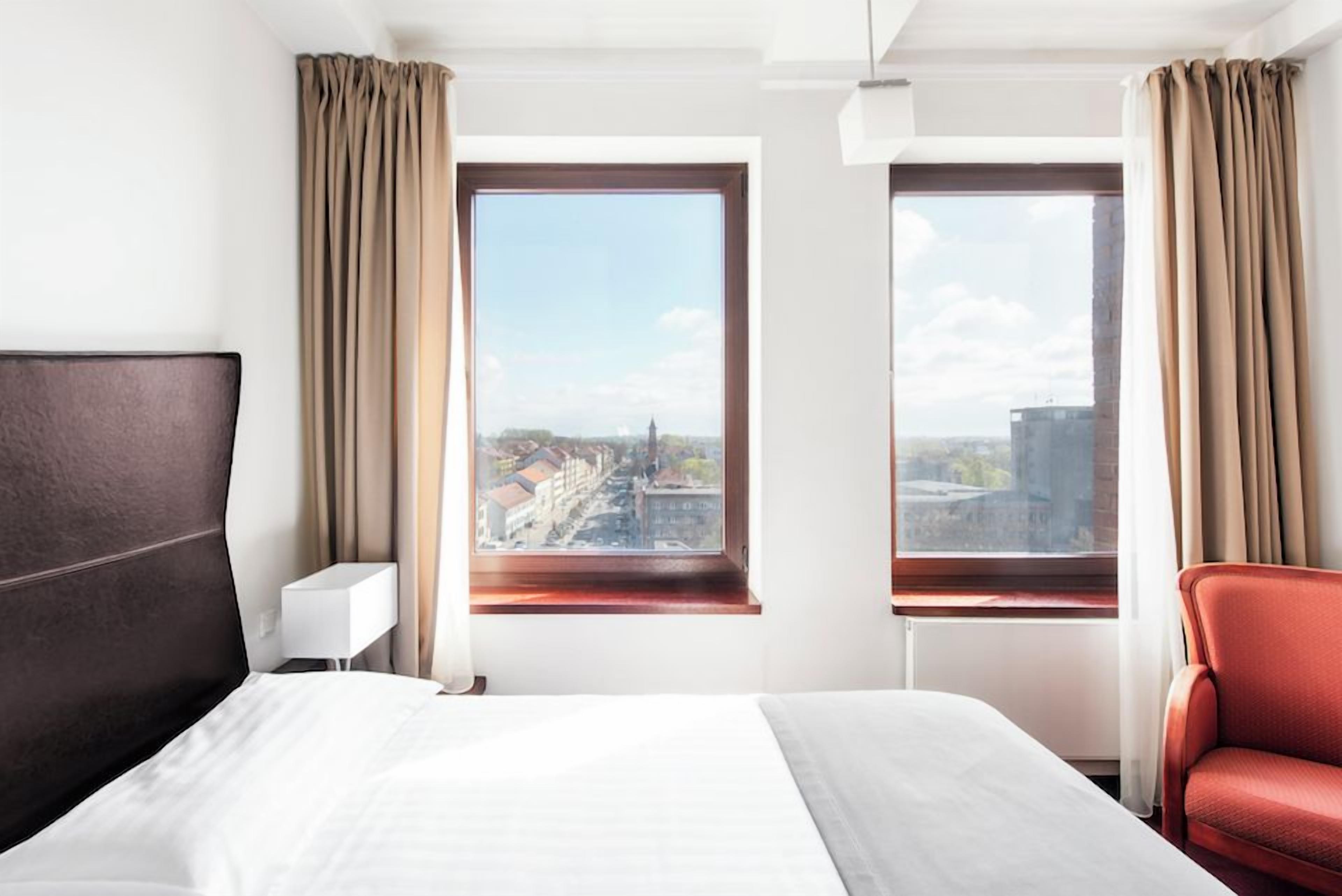 A hotel room with a large bed, two windows with curtains, a red chair, and a white bedside table.