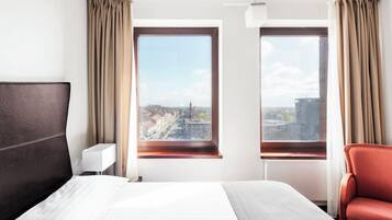 A hotel room with a large bed, two windows with curtains, a red chair, and a white bedside table.