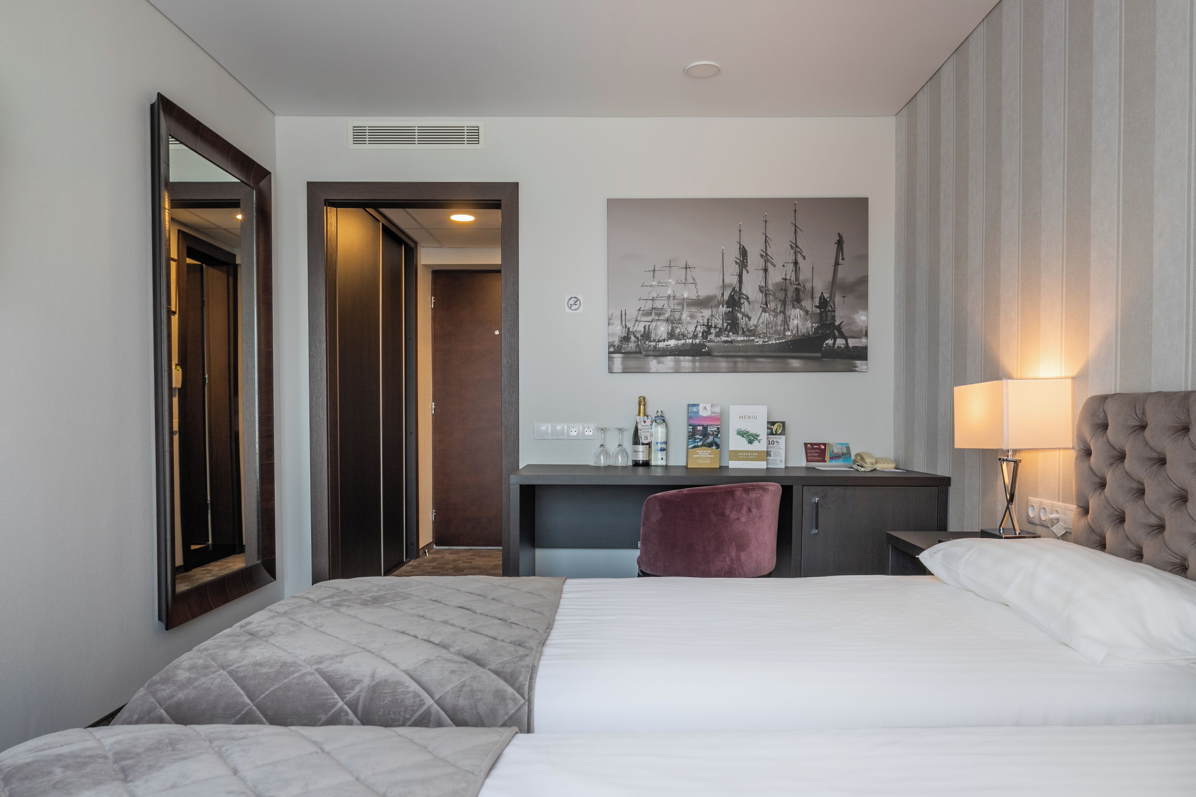 A hotel room with a bed, a desk, a chair, a mirror, a painting, and a lamp.