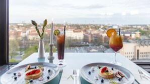 A table with two plates of food, two drinks, and a vase with flowers, set against a city view. - Amberton Hotel Klaipeda (Klaipeda)