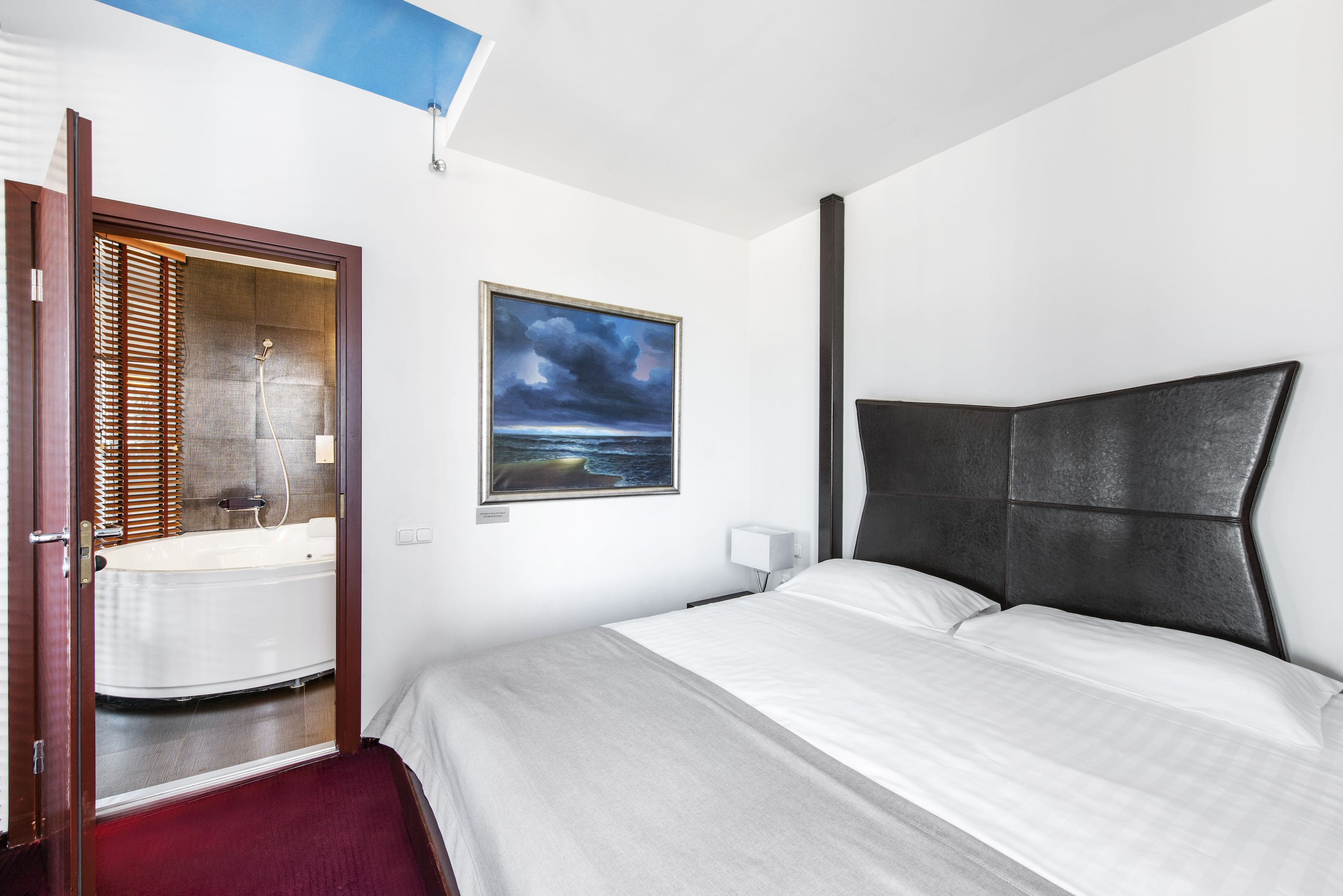 A hotel room with a bed, a painting of a beach scene, a bathtub, and a window with blinds.