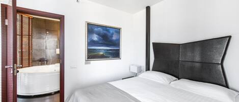A hotel room with a bed, a painting of a beach scene, a bathtub, and a window with blinds.